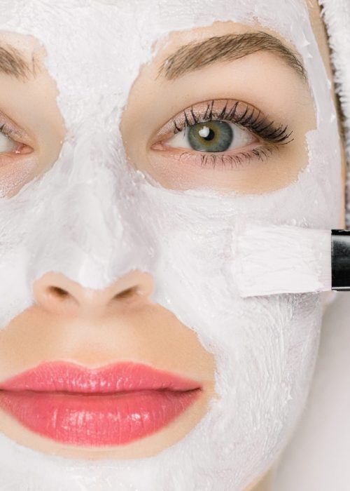 Facial beauty treatment. Closeup of pretty woman getting mask at spa salon. Cropped image of special brush in hands of cosmetologist applying white mud facial mask with on female face.
